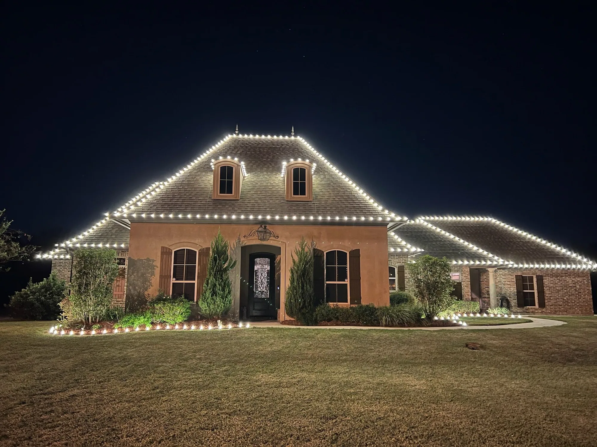 Brick Acadian home at night outlined in warm-white roofline lights with landscape uplighting