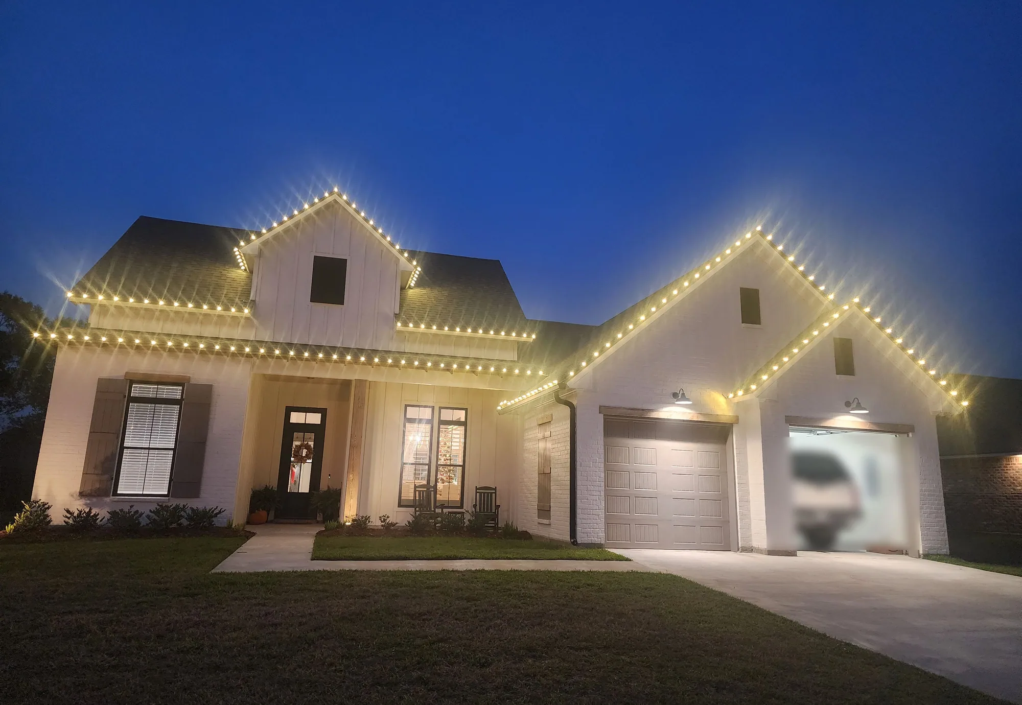 White modern farmhouse at twilight with warm-white Christmas lights outlining both rooflines and a lit front porch