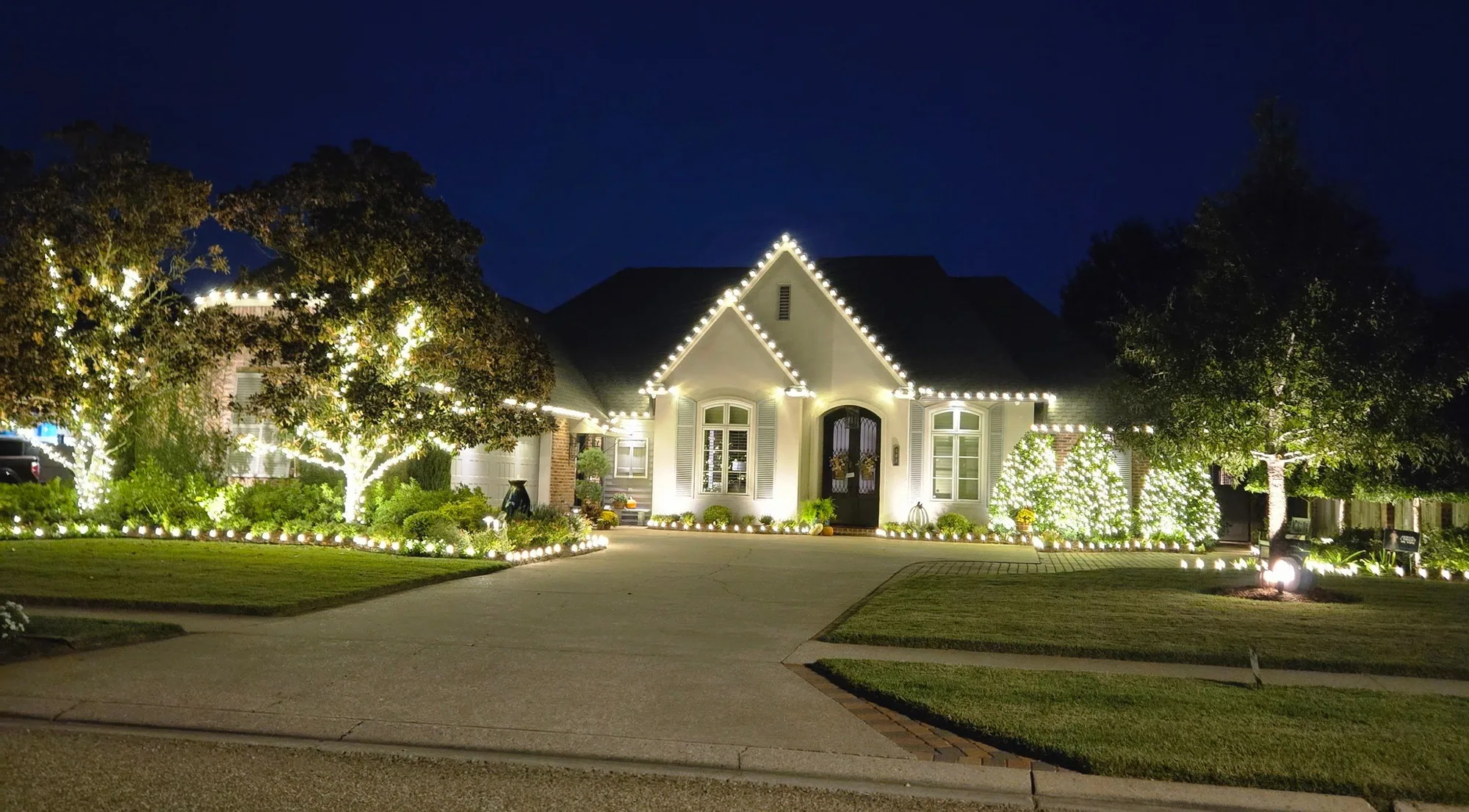 Two-story home at night with warm-white roofline lights, lit landscape trees and glowing entry