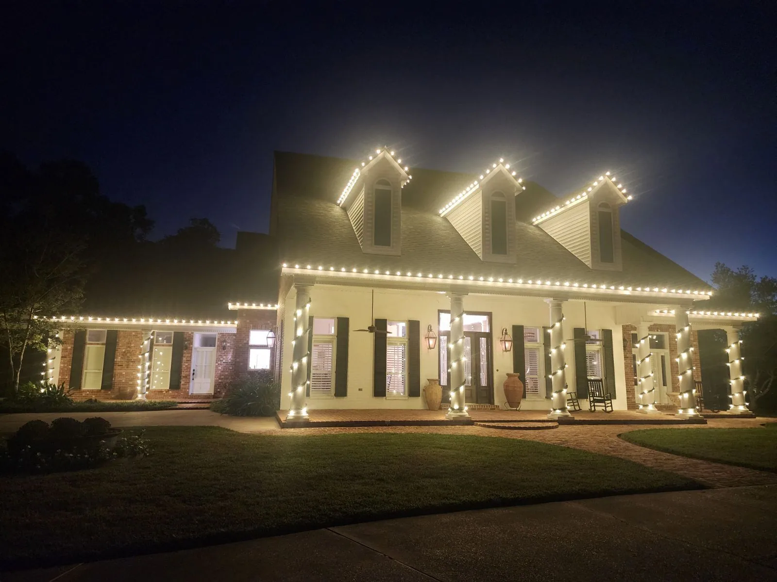 Large Acadian home at night with warm-white roofline lights and lit garland wrapping the porch columns
