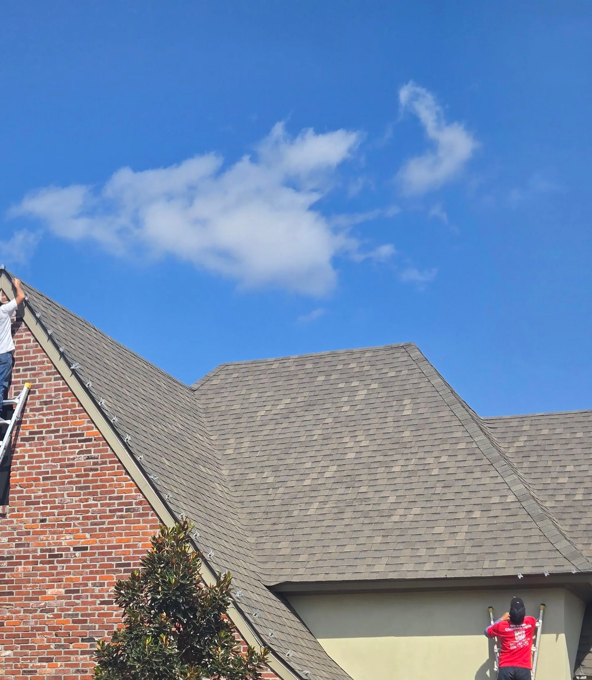 Two installers on ladders attaching warm-white Christmas lights along a home's roofline during the day
