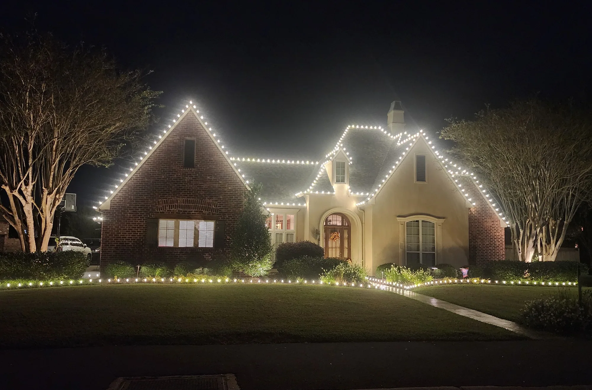Brick home at night with warm-white roofline lighting and landscape uplighting