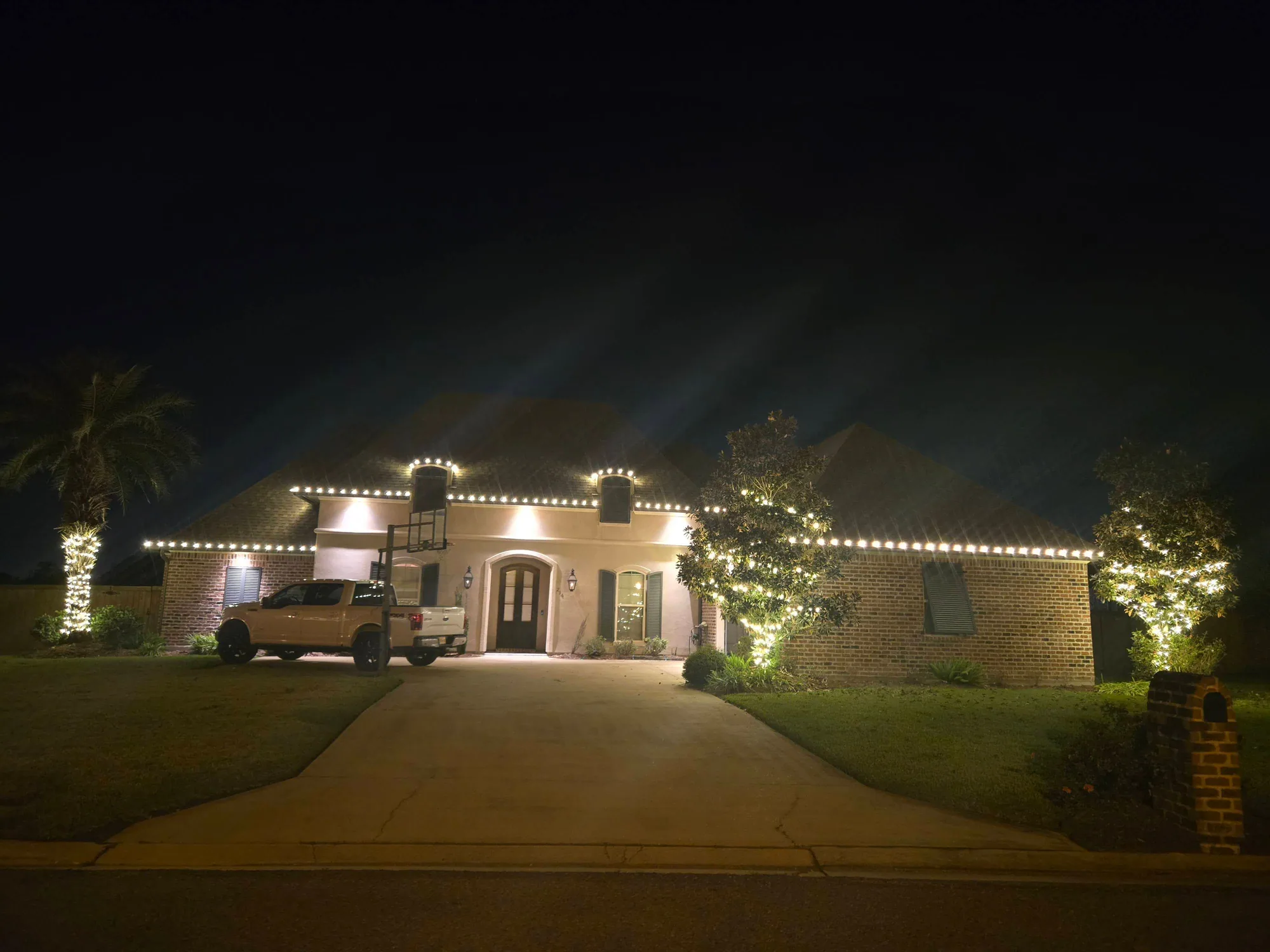Ranch home at night with warm-white roofline lights, a lit palm and landscape uplighting