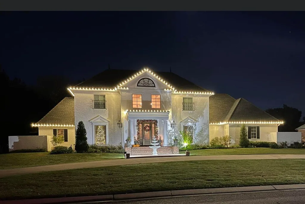 Two-story stucco home at night with warm-white roofline lights and a lit Christmas tree in the window