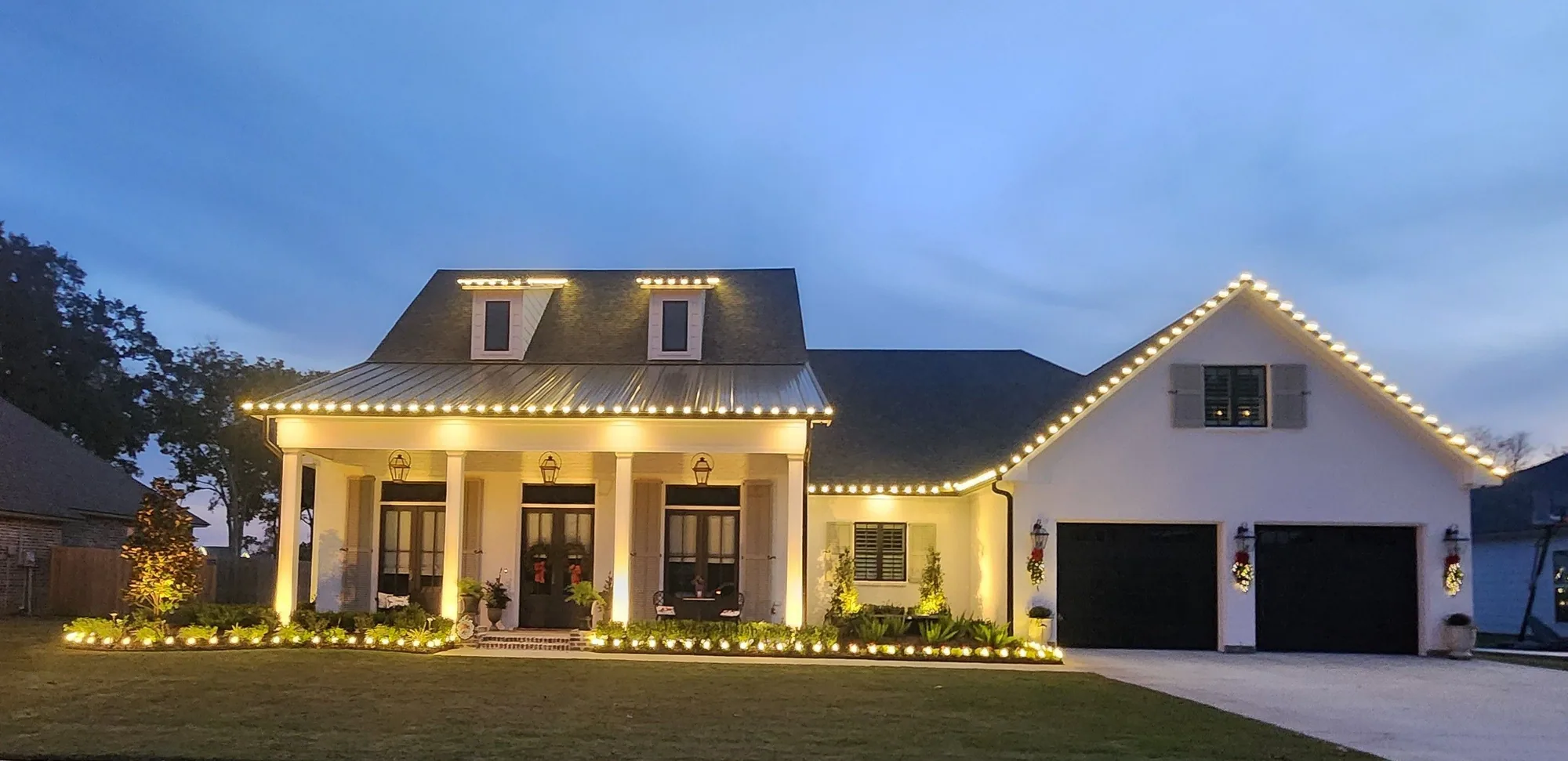 White farmhouse at twilight with warm-white roofline lights and lit porch columns