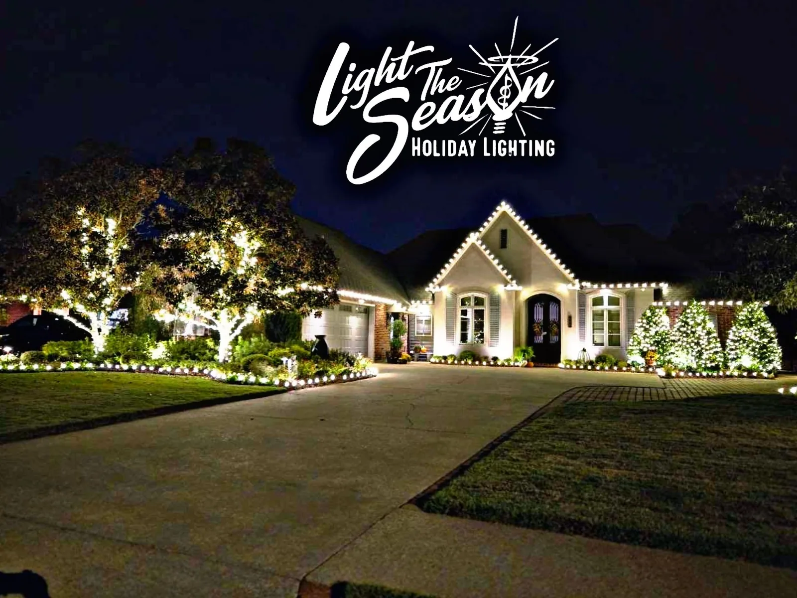 Acadian home at night with warm-white roofline lights and lit trees across the front yard