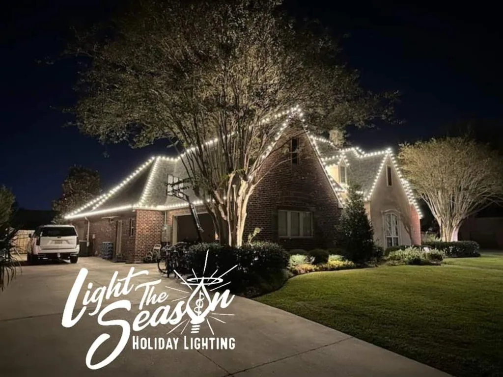 Large oak tree wrapped in warm-white lights beside a decorated home at night